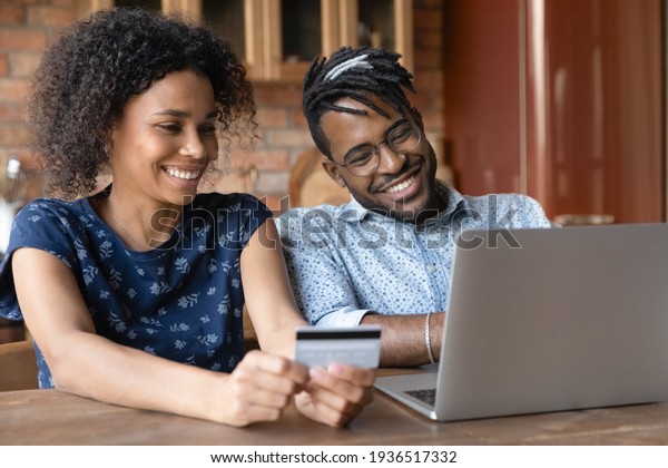 Paying online. Smiling young married couple of afro
american ethnicity provide payment via electronic bank app from
home office. Happy black spouses buy goods service at web use money
on prepaid card