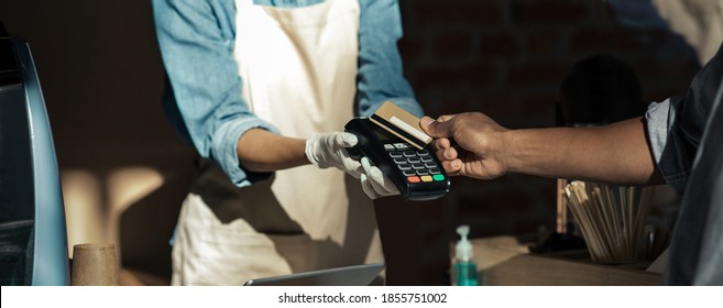 Payment for order and social distance during COVID-19 epidemic. Young african american barista in apron and gloves gives terminal for payment to client in modern cafe, cropped, panorama