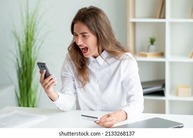 Payment Problem. Furious Woman. Mobile Application. Shouting Mad Office Lady Holding Credit Card Looking Smartphone Sitting Work Desk In Light Room Interior.