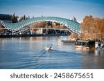  Peace Bridge, a modern architectural marvel, stretching across the Kura River in Tbilisi. The autumnal trees along the riverside add a seasonal charm, while boats navigate the tranquil waters, 