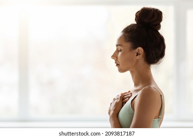 Peace Of Mind. Side View Shot Of Young Female With Hands On Chest And Closed Eyes, Profile Portrait Of Relaxed Woman Meditating Or Praying While Standing Against Big Window Indoors, Free Space