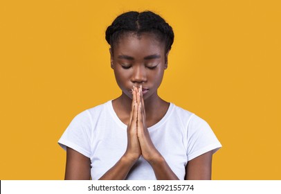 Peaceful African American Young Lady Praying Holding Clasped Hands In Prayer Gesture Standing With Eyes Closed Posing In Studio Over Yellow Background. Portrait Of Black Woman Sending Prayers To God