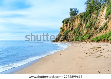 Peaceful atmosphere of a an ocean coastline in Malibu, California. High and lush green cliff towers above the sandy beach, calm ocean waters stretch out to the horizon under a clear blue sky.