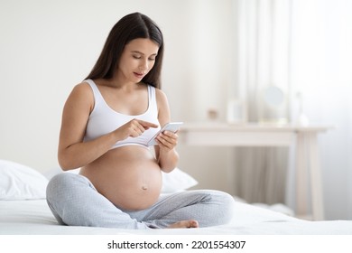 Peaceful attractive young expecting lady sitting on bed, using cell phone, touching gadget screen and smiling, shopping on Internet, using mobile app for pregnant women, bedroom interior, copy space - Powered by Shutterstock