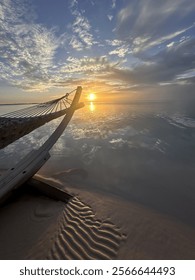 A peaceful beach scene with a hammock gently swaying as the sun sets, casting a warm glow across the water and reflecting in the calm, serene sea.