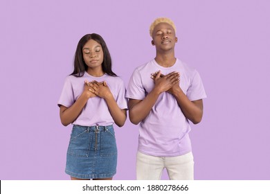 Peaceful Black Couple Putting Hands On Heart, Praying To God With Closed Eyes Over Violet Studio Background. Young Woman And Her Boyfriend Cherishing Hope For Best Or Meditating