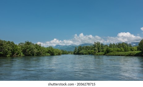 A peaceful blue river flows between the banks with lush green vegetation. In the distance, against the background of the azure sky and clouds, a mountain range is visible. Kamchatka