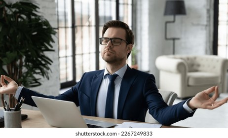 Peaceful business leader meditating at workplace, making zen hands and fingers at laptop. Calm businessman relaxing in office, breathing, doing yoga, practicing mindfulness and stress relief exercises - Powered by Shutterstock