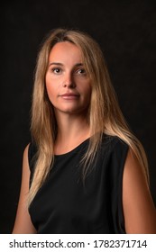 Peaceful Calm Beautiful Girl In A Black Dress On A Dark Background, Portrait Of A 30 Year Old Woman With Long Blonde Hair. Vertical Sad Studio Shot.