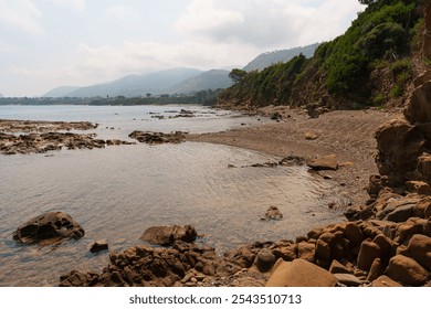 A peaceful coastal beach scene featuring a rocky shoreline, calm waters, and distant hills. The natural landscape provides a serene environment for relaxation and exploration - Powered by Shutterstock