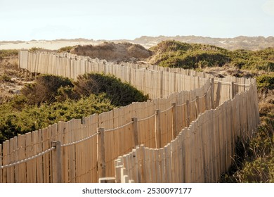 A peaceful coastal pathway bordered by rustic wooden fencing, winding through sandy dunes under a warm, sunny sky, offering a tranquil and scenic outdoor experience. - Powered by Shutterstock