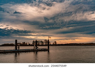 A peaceful dock scene at sunset with a lone flag waving under a dramatic, cloudy sky. Calm waters and warm hues create a tranquil atmosphere, capturing the beauty of nature and stillness - Powered by Shutterstock