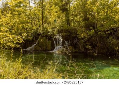 A peaceful forest scene featuring a small waterfall flowing into a clear, green water pool surrounded by lush greenery, in Croatia - Powered by Shutterstock