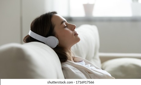 Joven tranquila con sus modernos auriculares inalámbricos sentada relajada en un cómodo sofá escuchando música, joven feliz y tranquilo en auriculares descansa en un acogedor sofá, disfruta de sonido de buena calidad, concepto sin estrés