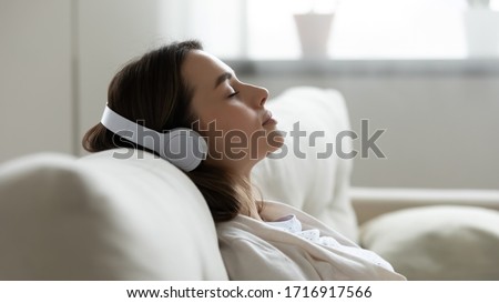 Peaceful girl in modern wireless headphones sit relax on comfortable couch listening to music, happy calm young woman in earphones rest on cozy sofa, enjoy good quality sound, stress free concept