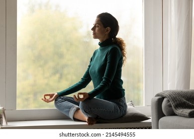 Peaceful Hispanic Woman Sit On Windowsill At Home Practice Yoga With Mudra Hands Breathe Fresh Air. Calm Latino Female Meditate Indoors Relieve Negative Emotions. Peace, Stress Free Concept.