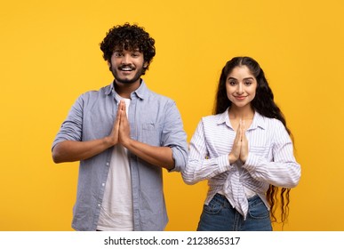 Peaceful indian couple putting hands together in namaste or prayer gesture, looking at camera while posing on yellow studio background