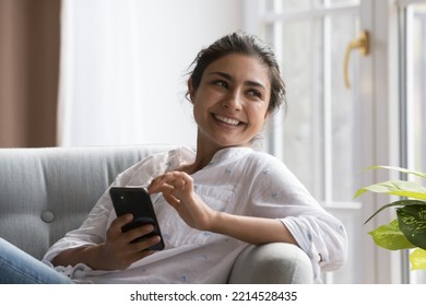 Peaceful Indian woman relaxing on cozy chair with smartphone, smile staring aside enjoy carefree weekend and modern wireless tech. Making call, ordering goods and services on-line, e-date apps usage - Powered by Shutterstock