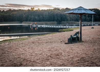 A peaceful lakeside landscape at sunset with a sandy beach with an empty wooden bench under a thatched roof. A pier with a horizon of trees stretches out into the calm water. - Powered by Shutterstock