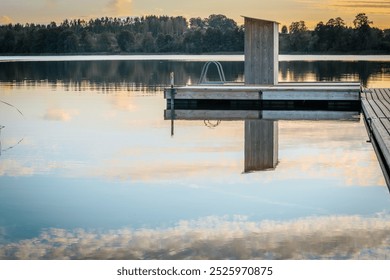 A peaceful lakeside landscape with a wooden pier stretching out into the calm water and reflecting the golden hues of the sunset. A small staircase and a floating platform add to the peaceful atmosphe - Powered by Shutterstock