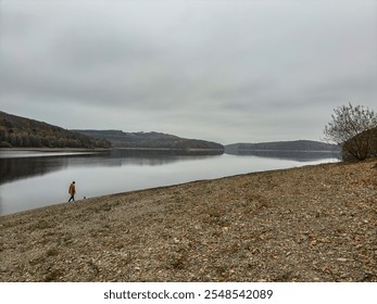 A peaceful lakeside scene on a cloudy day, featuring a lone person in a yellow jacket walking along the rocky shore. Calm water reflects the overcast sky and surrounding forested hills. - Powered by Shutterstock