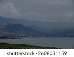 A peaceful lakeside village lies at the foot of majestic mountains, shrouded in a blanket of gray clouds as evening descends. The quiet waters reflect the tranquil beauty of nature.