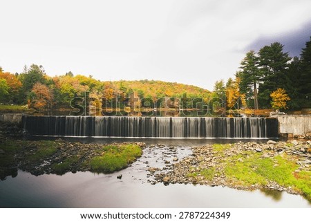 Peaceful landscape featuring a smooth waterfall cascading over a spillway, framed by trees, calm water, and a serene natural setting with vibrant outdoor scenery.