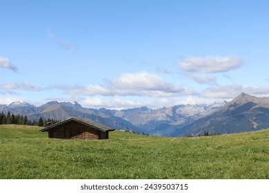 peaceful landscape with green mountain meadow, brown wooden hut and blue mountain range and sky - Powered by Shutterstock