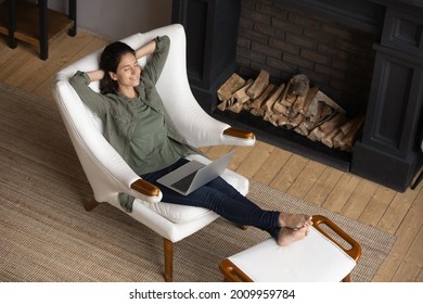 Peaceful latina woman lean back in comfy chair with hands over head take break in work with laptop relax at modern living room near fireplace. Calm young lady enjoy rest at home breath clean fresh air