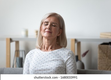 Peaceful middle aged blonde woman leaning on sofa, relaxing with closed eyes. Calm mature retired lady resting in silence, meditating, visualizing future, enjoying weekend leisure time alone at home.