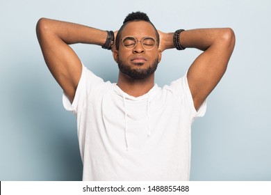 Peaceful millennial african American man isolated on blue studio background relaxing hands over head, calm black male with eyes closed rest having nap or fall asleep standing. Stress free concept