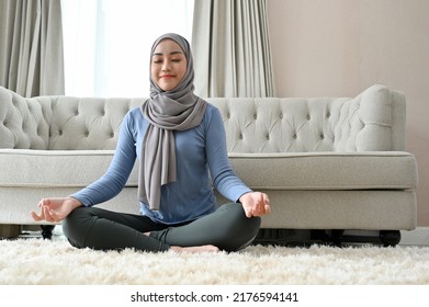 Peaceful millennial Asian Muslim woman with hijab practicing yoga, concentrated meditating, doing a lotus pose in the living room. - Powered by Shutterstock