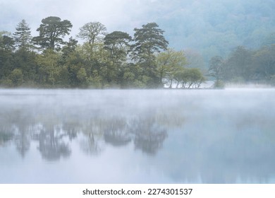 Peaceful morning at lake with mist rising from water and calm reflections of trees. Rydal Water, Lake District, UK. - Powered by Shutterstock