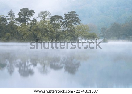 Peaceful morning at lake with mist rising from water and calm reflections of trees. Rydal Water, Lake District, UK.