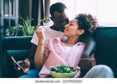 Peaceful multiethnic people in relationship kissing around with each other while eating salad and using smartphone on weekend at home - Powered by Shutterstock