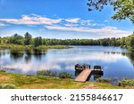 A peaceful Northern Wisconsin forest lake with a wooden pier reflects the blue sky and white clouds of a summer day amid lush green marshes and forestland.