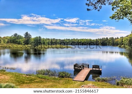 A peaceful Northern Wisconsin forest lake with a wooden pier reflects the blue sky and white clouds of a summer day amid lush green marshes and forestland.