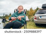 Peaceful Norwegian camping scene with teen boy playing ukulele surrounded by nature by lakeside. relaxing in chair while camp days with car in wilderness.