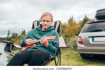 Peaceful Norwegian camping scene with teen boy playing ukulele surrounded by nature by lakeside. relaxing in chair while camp days with car in wilderness. - Powered by Shutterstock