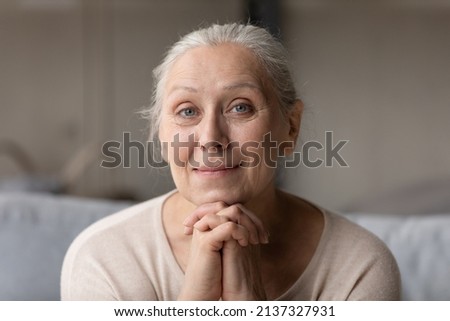 Peaceful older woman smile looks at camera, head shot portrait. Attractive granny put chin on folded hands, relax alone on sofa at home. Retirement, medical insurance cover for elderly citizen concept