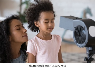 Peaceful Overheated African Mom And Little Daughter Kid Facing Cool Fresh Air Flow From Electric Fan, Blower, Cooler, Ventilator. Home Appliance, Summer Weather, Comfortable Family House Concept