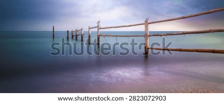 Peaceful panoramic seascape featuring a rustic wooden pier or fence extending into calm ocean water, with a seagull perched on a post near the horizon. The image is captured from a low coastal viewpoint with a wide horizontal composition, soft long exposure water, muted turquoise and blue tones, and a cloudy dramatic sky. The minimalist marine landscape creates a serene, tranquil, and contemplative mood, ideal for travel advertising, nature backgrounds, coastal tourism, wellness themes, relaxation concepts, environmental design, editorial layouts, website banners, and scenic wall art.
