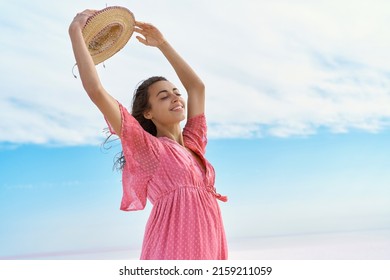 Peaceful portrait of carefree pleased woman in pink dress holding hat over head on blue sky background