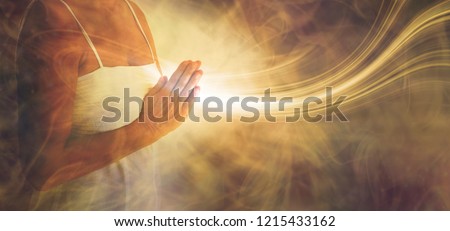 Peaceful prayer sending love and light out -  female in white dress with hands in prayer position and a stream or white light flowing outwards with a rustic golden brown ethereal energy background
