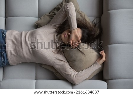Peaceful pretty 20s girl lying on back on comfortable couch at home, covering eyes with hand, sleeping at daytime, feeling tired, enjoying relaxation, leisure, break, weekend. Top view