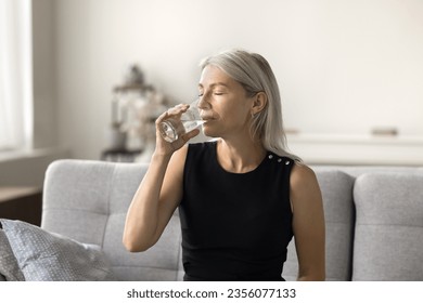 Peaceful pretty blonde mature woman drinking natural mineral water with closed eyes for healthcare, healthy metabolism, refreshment, sitting on couch at home, satisfying thirst - Powered by Shutterstock