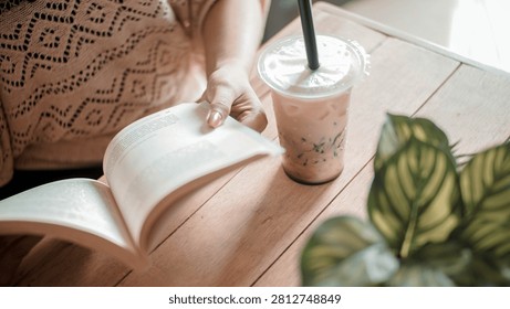 A peaceful reading setup featuring an open book, iced coffee, and decorative green houseplant on a wooden table. The image highlights leisure, learning, mindfulness, and slow living concepts.
