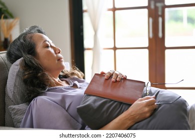 Peaceful And Relaxed Aged Asian Woman Fall Asleep While Reading A Book On Her Comfortable Sofa In Her Living Room. Retired Woman's Lifestyle Concept