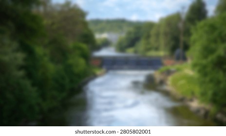 A peaceful river flows through a dense green forest under a cloudy sky with a small weir structure visible in the distance
