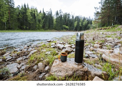 At a peaceful riverbank, a sleek stainlesssteel thermos and an attractive mug sit on a smooth rock, surrounded by lush greenery and flowing water, perfect for outdoor adventures and relaxation